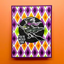 A handmade greeting card featuring a witch riding a broomstick over a black circle with the text 'YOU'RE WICKED COOL', set against a purple, orange, and white harlequin diamond pattern. The card is displayed flat on a solid orange background.
