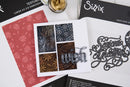 A flat-lay of crafting supplies showing a white card with four decorative panels and the word 'wish' in 3D, alongside patterned embossing papers, a gold foil sheet, and black cards branded Sizzix.