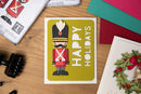 A handmade holiday card featuring a nutcracker soldier and the words 'HAPPY HOLIDAYS' on a yellow-green background, styled on a wooden table with crafting supplies.