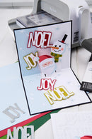 A handmade 3D pop-up Christmas greeting card displayed in a crafting workspace. The card features a Santa Claus and a snowman with layered text reading 'NOEL JOY NOEL' in red, white, and gold.
