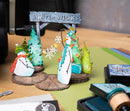 A handmade winter-themed paper craft display featuring two snowman figures, decorated pine trees, a 'Winter Wishes' sign, and gift boxes on a wooden surface, with ink pads nearby.