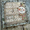 A rustic decorative wooden sign with the phrase "let it snow let it snow let it snow" in distressed white lettering, featuring a raised snowman face and scarf on the right. The sign has a distressed metal border and is photographed on weathered wood planks.