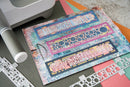 A close-up of a crafting sheet with colorful abstract rectangular panels and decorative cutout pieces, placed on a work surface with a white machine and metal dies visible around it.
