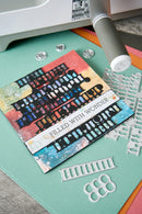 A close-up of a handmade greeting card with a colorful architectural illustration and the text 'FILLED WITH WONDER,' shown alongside metal die-cutting and scoring tools on a teal crafting mat.