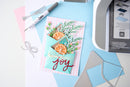A handmade layered card with teal envelope details, green foliage, orange slices, and the word 'joy' in red script, styled on a light blue and white surface with crafting tools nearby.