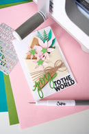 A handmade greeting card with layered paper flowers, green leaves, and the text 'you TO THE WORLD' in green and black, styled on a pink surface with crafting tools nearby.