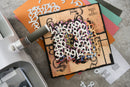 A square craft die-cut paper design featuring the number 1 in white with layered multicolored numerals and decorative black swirls on a tan background. The piece is displayed on top of orange and pastel cardstock with a die-cutting machine partially visible on the left.