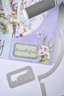 A close-up of a lavender scrapbooking paper sheet with floral and butterfly illustrations, a decorative label reading 'beautiful,' and silver metal die-cutting or stamping tools placed on top and around it.