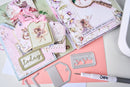 A styled flat-lay of scrapbooking and crafting supplies, including layered paper albums with fairy and floral designs, metal die-cutting templates, a pink pen, and a white cutting machine on a light gray surface.