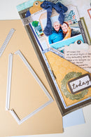 A close-up of a scrapbook or card-making craft kit showing layered paper elements, a photo of two people, decorative tags, a blue ribbon, and metal die-cut frames on a beige background.