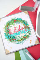 A square handmade greeting card with a festive wreath design and the words 'holiday memories' in pink and teal script, styled on a red background with crafting tools partially visible.