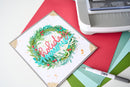 A square handmade greeting card with a festive wreath design and the words 'holiday memories' in layered script, placed on top of red and green cardstock with a white die-cutting or stamping machine partially visible in the upper right.