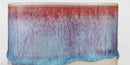 A close-up of a ceramic planter or vase with a dripping glaze finish, featuring a gradient transition from reddish-brown at the top to light blue at the bottom, set against a plain white background.