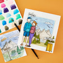 Two illustrated cards or prints laid on an orange surface, one showing a couple with mountains and the other a simple congratulations scene with mountains. A paint palette sheet, paintbrush, and yellow marker are arranged around them as styled props.
