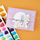 A handmade greeting card or craft paper piece on a solid orange background, styled with a watercolor paint palette and paintbrushes. The card is lavender purple with a grid texture, featuring a central white oval with a cartoon girl, the phrase 'it's your day' in black script, and layered pink and green paper flowers.