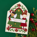 A handmade Christmas angel card featuring two paper-cut angels with wings, set against a green background with holiday decorations including red berries and evergreen branches.
