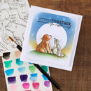 Card with two bears sitting together, surrounded by watercolor swatches and a paintbrush on a wooden surface.