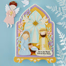 A decorative Christmas-style nativity scene plaque or card featuring layered paper-cut figures of an angel, Mary, Joseph, and baby Jesus in front of a stained-glass-style window with doves. The plaque has a pastel color palette and the text 'Joy to the World Love to Your Home' at the bottom.