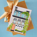 A handmade greeting card featuring a detailed illustration of a house with black-and-white awnings, green ivy, and the text 'A Happy little House from my house to yours,' styled on top of a kraft envelope with a gold ribbon against a light blue background.