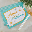 A handmade holiday greeting card with 'Happy Holidays' in gold and teal script, decorated with two white daisy flowers, shown on a turquoise gingham background next to a white and gold pen.