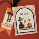A Halloween-themed greeting card and matching tag on a warm orange-brown background. The main card has a gingham border and an arched illustration of three cartoon geese dressed as witch, ghost, and pumpkin.