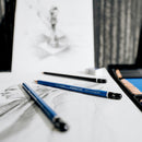 Staedtler drawing pencils on a white surface with blurred artwork in the background