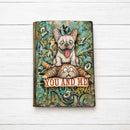 A hardcover notebook or journal featuring a whimsical illustration of a French bulldog sitting on a cat with the text 'YOU AND ME' across the front. The cover has a distressed, vintage-style teal and brown background with scattered numbers.