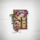 A decorative spiral-bound notebook with a whimsical pop-art design featuring cartoon cats, a purple 'POP' title, and small bottle and ribbon embellishments, photographed on a white wood background.