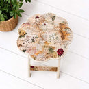 A small scalloped side table with a vintage-style floral and script print on the tabletop and a matching patterned wooden base, photographed on a white wood plank floor next to a potted green plant.