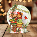 A round Christmas-themed decorative plaque or coaster featuring two kissing mice, wrapped gifts, red hearts, holly, and the text 'MERRY KISS-MOUSE' on a wooden tabletop with warm blurred lights in the background.
