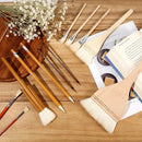 A set of wooden-handled art brushes and a large palette brush arranged on a wooden surface with flowers, an open book, and paper props.