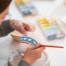 A close-up of a person's hands painting a white ceramic mug with a blue bridge design and a red paintbrush, set in a bright pottery studio with blurred paint palettes in the background.