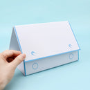 A hand holding a white and light blue house-shaped desk organizer with two circular cutouts on the front, set against a solid light blue background.