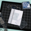 A clear acrylic stamp block with the words 'WISH YOU WELL' in raised letters, placed on a white sheet of paper over a black gridded cutting mat. Surrounding items include a clear acrylic ruler, a small square blue-and-white printed stamp or ink pad, and additional clear stamps with decorative text.