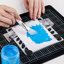 A close-up of hands mixing bright blue gel or paint on a white sheet over a black grid cutting mat, with a small blue jar in the foreground.