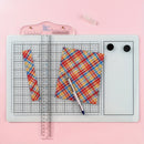 A top-down view of a white and gray grid cutting mat on a pink background, featuring a pink plastic ruler, a plaid patterned paper sheet, a white pen, and a small clear accessory.