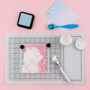 A flat-lay of a craft stamping kit on a pink background, featuring a white floral stamp on a pink block, a gray gridded cutting mat, a blue ink pad, a blue brush, and small white containers of wax or paste.