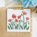 A square white card or small sign clipped at the top with a gold binder clip, featuring embroidered poppy flowers in pink, orange, and green on a burlap surface with decorative fabric and flowers around it.