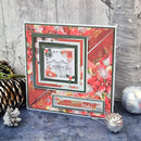 A handmade square Christmas greeting card with layered floral and geometric patterns in red, green, and white, featuring a small central illustration of a house. The card is styled on a dark textured surface with holiday ornaments and a pinecone against a gray wood-plank background.