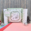 A handmade layered Christmas pop-up card with vintage-style details, a rustic wood background and holiday props including a small tree, pinecone, and ornament.