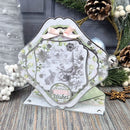 A decorative handmade greeting card with layered scalloped edges, a pale pink bow, and a central winter scene featuring a gnome, squirrel, snowflakes, and the words 'Warmest Winter Wishes.' The card is styled on a rustic wood surface with pine branches, pinecones, white berries, and holiday ornaments in the background.
