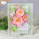 A handmade greeting card featuring layered pink and peach paper flowers with green leaves on a textured pale green background, displayed on a white lace surface.