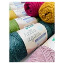 A close-up arrangement of Grounds 'hello again' wool yarn balls in multiple colors, including teal, pink, mustard yellow, light green, and dusty pink, with paper labels visible.