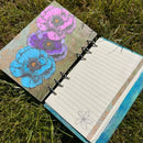 An open ring-bound notebook lying on green grass, showing lined pages on one side and a decorated cover on the other with three large poppy flowers in blue, purple, and pink over a textured beige background.