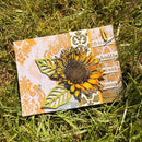A handmade mixed-media card featuring a large layered sunflower, green leaf cutouts, vintage-style orange and white damask patterns, and text strips, photographed lying on green grass.
