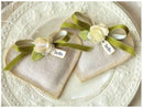 Two beige fabric napkins tied with olive green ribbons and decorated with cream fabric flowers, displayed on a white textured ceramic plate.