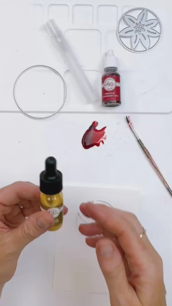Hands holding a small amber dropper bottle filled with yellow liquid, shown alongside other craft or dyeing tools on a white surface, including a small red bottle, a metal ring, a clear tube, a paintbrush, and a red liquid swatch.