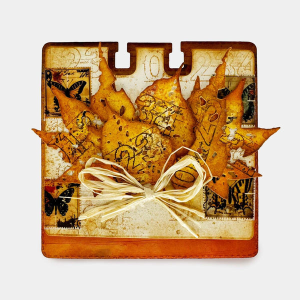 A square decorative card or art piece featuring layered autumn leaves in warm orange, yellow, and brown tones, with vintage-style text, butterfly motifs, and a cream twine bow at the center, shown on a light gray background.