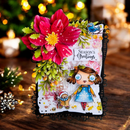 A handmade Christmas greeting card with a large pink flower, green leaves, a blue bird, and a cartoon girl holding a gingerbread cookie, displayed on a wooden surface with blurred holiday lights in the background.
