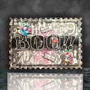 A square mixed-media art card featuring the word 'BOO!!' in large distressed black letters, decorated with colorful whimsical insect and bird illustrations, torn paper labels, and a gray serrated border, shown against a dark textured background with a reflection below.